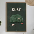 a framed poster with the word "BUSY" on it, hanging on a wall above a dining table with various items on it, such as a bottle, a bowl, and a vase.