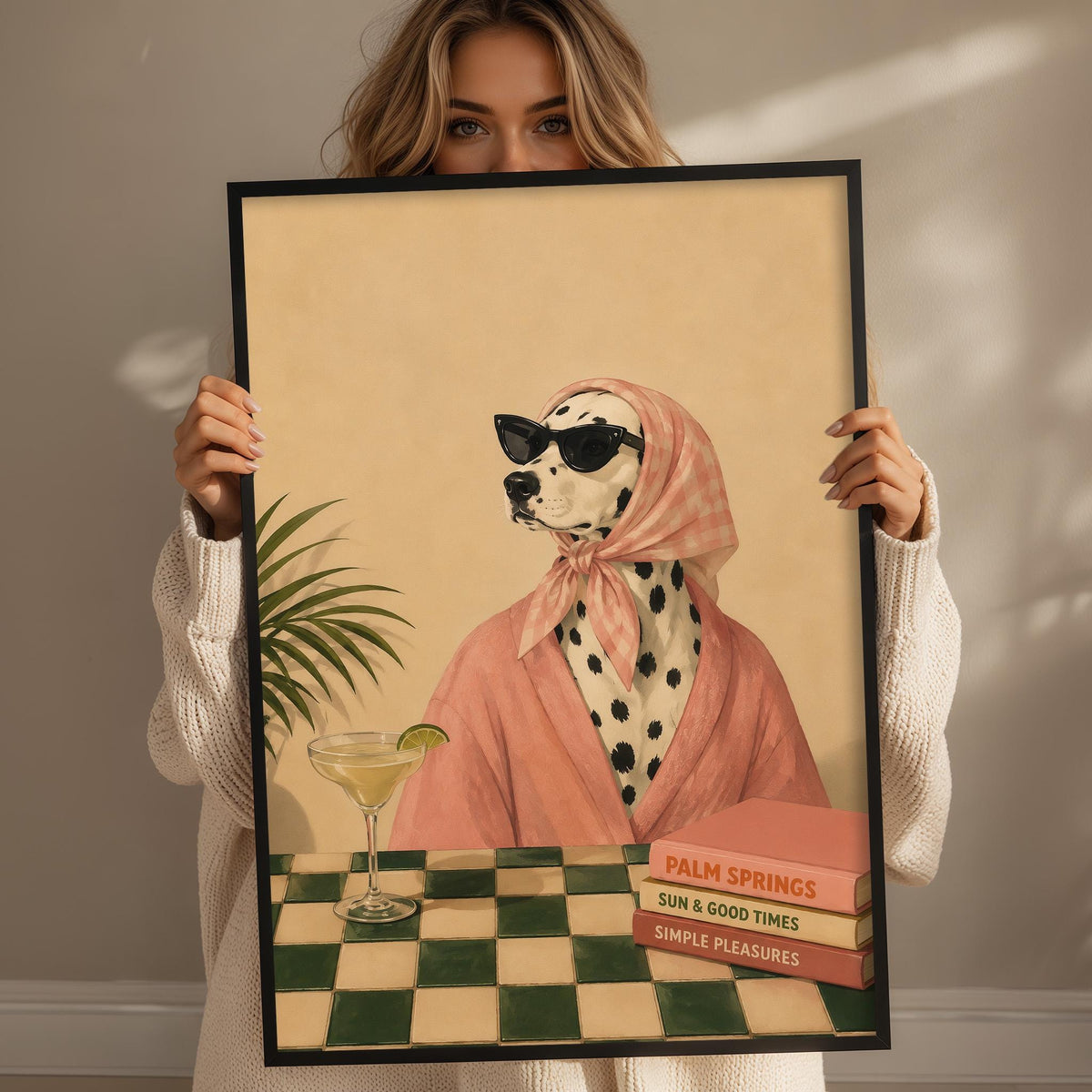 A woman holds up a framed print of a Dalmatian dog wearing a pink robe and sunglasses, sitting at a table with a drink and a book titled "Palm Springs".