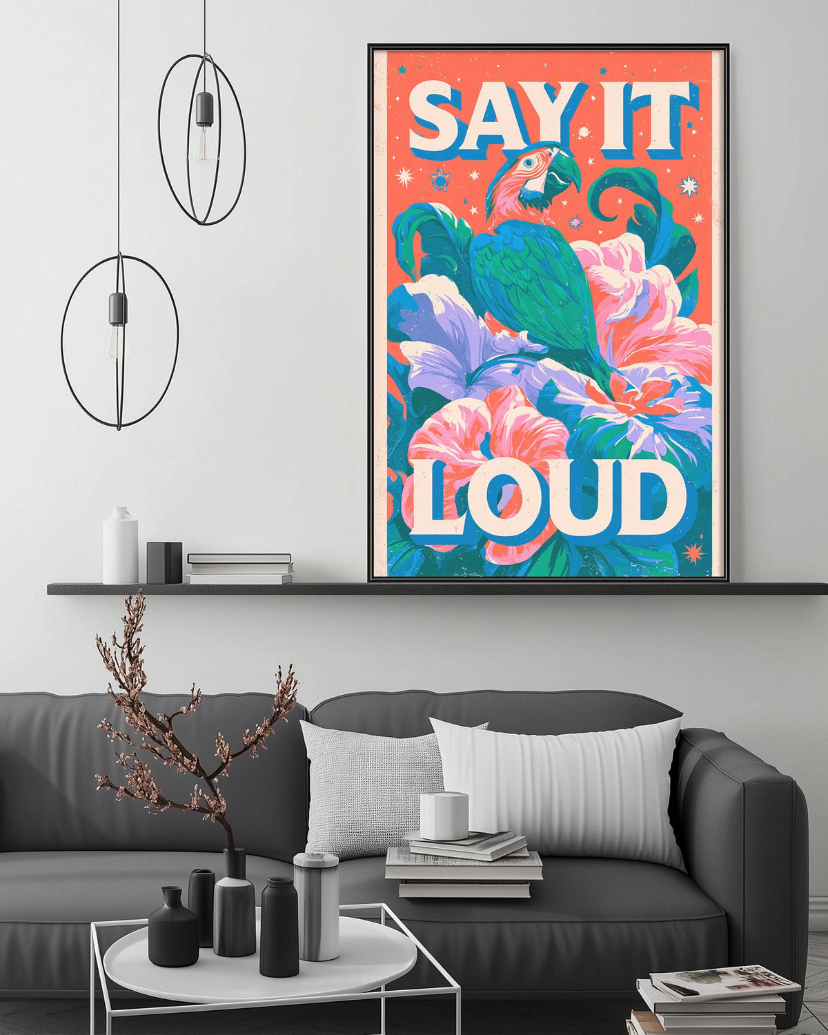 a large, colorful poster with the text "Say it loud" displayed prominently. The poster is hanging on a wall above a gray couch, which is positioned in front of a coffee table. The room has a minimalist and modern design, with a few decorative items such as vases and a potted plant.