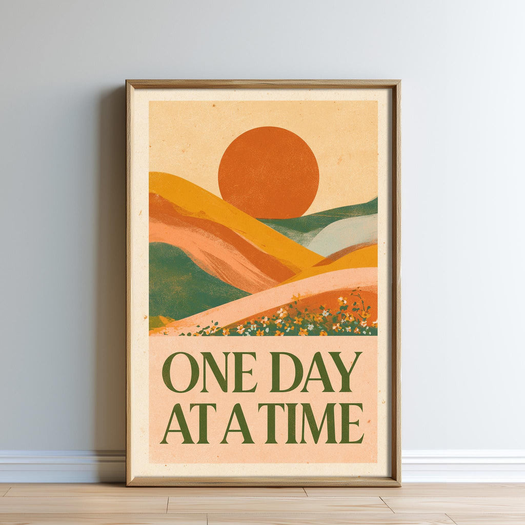 The image is a framed poster with a landscape scene featuring mountains, hills, and a large sun in the sky. The text on the poster reads "ONE DAY AT A TIME".