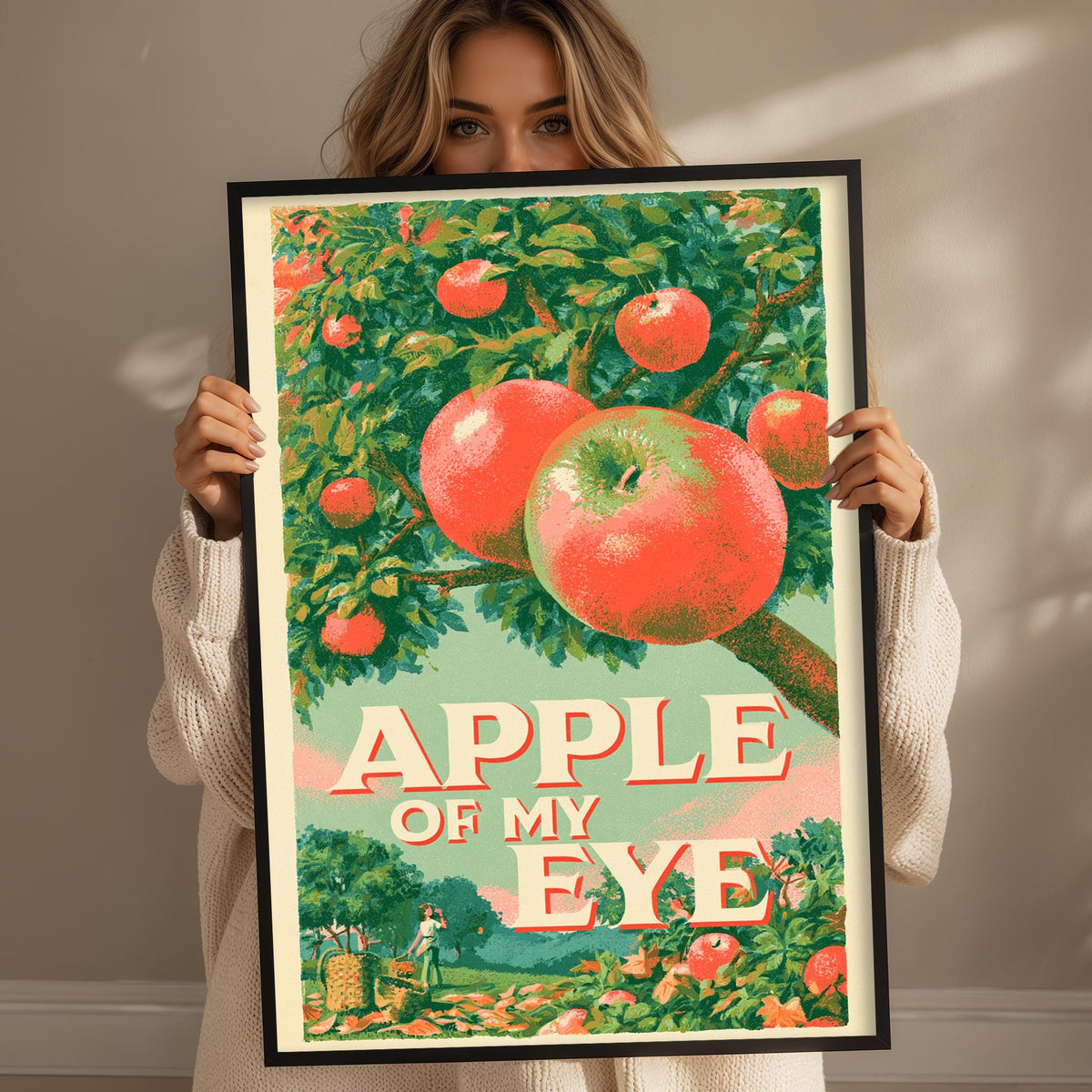 A woman holds up a framed poster with a vibrant illustration of an apple tree and the text "APPLE OF MY EYE" on it.
