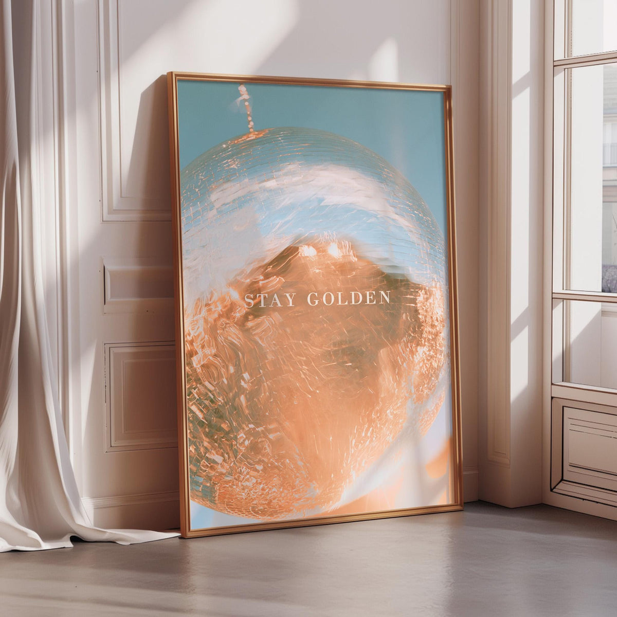 A framed poster with a vibrant, abstract design of a globe and the text "STAY GOLDEN" is displayed on a wall in a room with sunlight streaming in through a window.