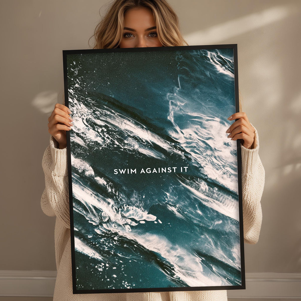 A woman holding a framed poster with a large ocean wave design and the text "SWIM AGAINST IT" printed on it.