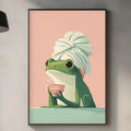 a framed illustration of a frog wearing a towel, sitting on a table and holding a cup. The background of the image is pink, and the frog is wearing a green hat. The kitchen counter is visible in the foreground, with various kitchen items such as a cutting board, a bowl, and a bottle.