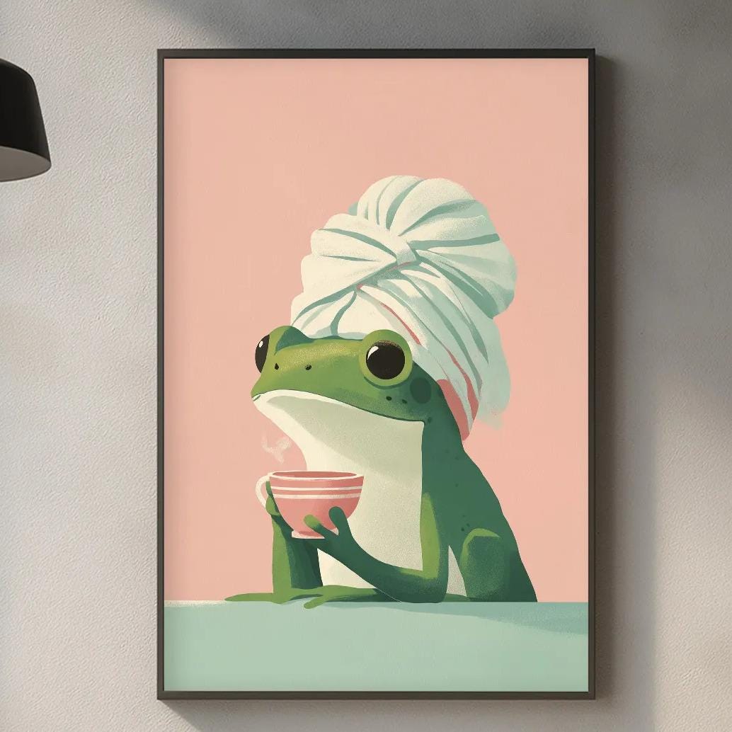 a framed illustration of a frog wearing a towel, sitting on a table and holding a cup. The background of the image is pink, and the frog is wearing a green hat. The kitchen counter is visible in the foreground, with various kitchen items such as a cutting board, a bowl, and a bottle.
