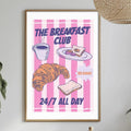 a poster for "The Breakfast Club" with illustrations of breakfast items such as a croissant, toast, and coffee. The poster is hanging on a wall above a dining table with various items on it, including a bottle, a bowl, and a vase.