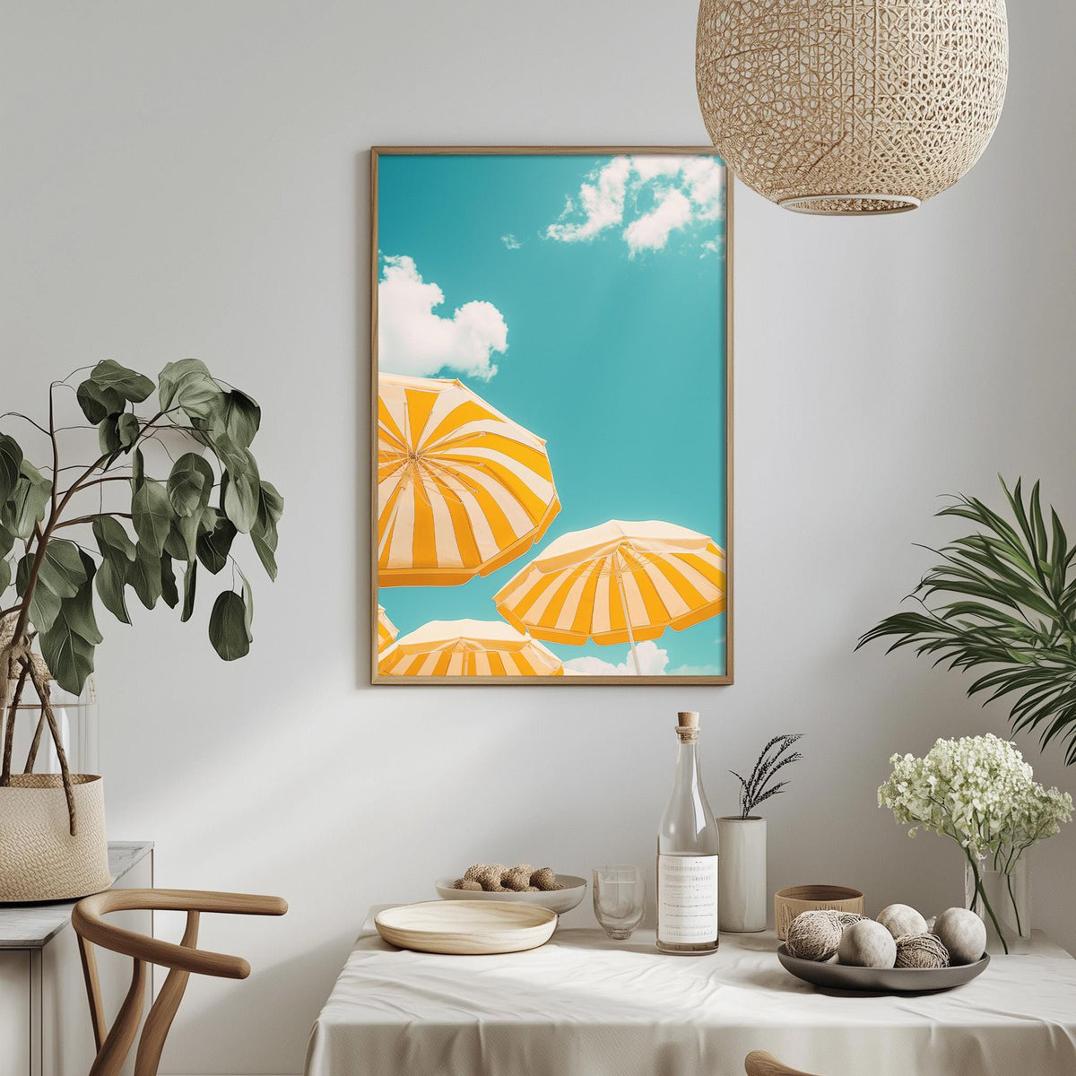 a dining table with a white tablecloth, a bowl of fruit, a bottle, and a vase with flowers. Above the table, there is a framed picture of three umbrellas against a blue sky with clouds.