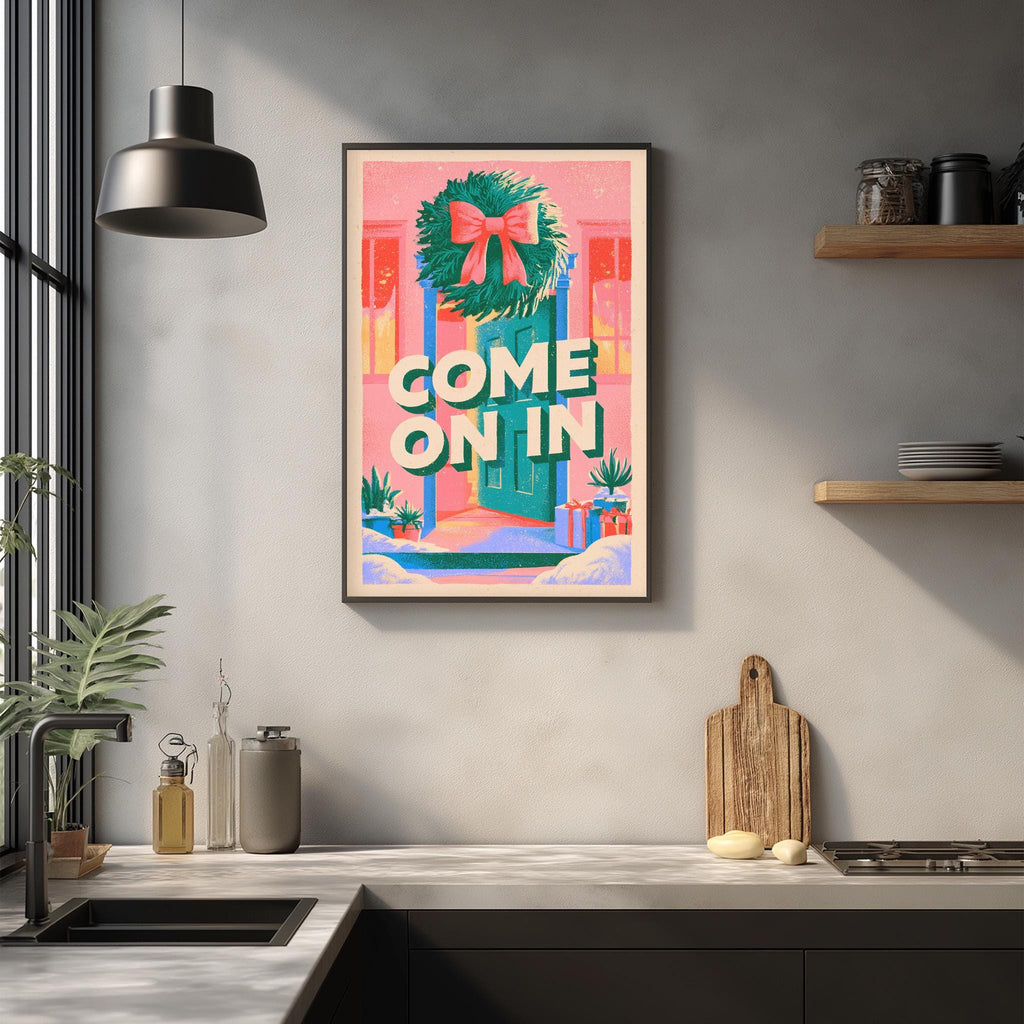 a kitchen counter with a framed poster hanging on the wall above it. The poster has a colorful design with a wreath and the text "COME ON IN" written on it. The kitchen counter has various items on it, including a cutting board, a bottle, and a bowl.