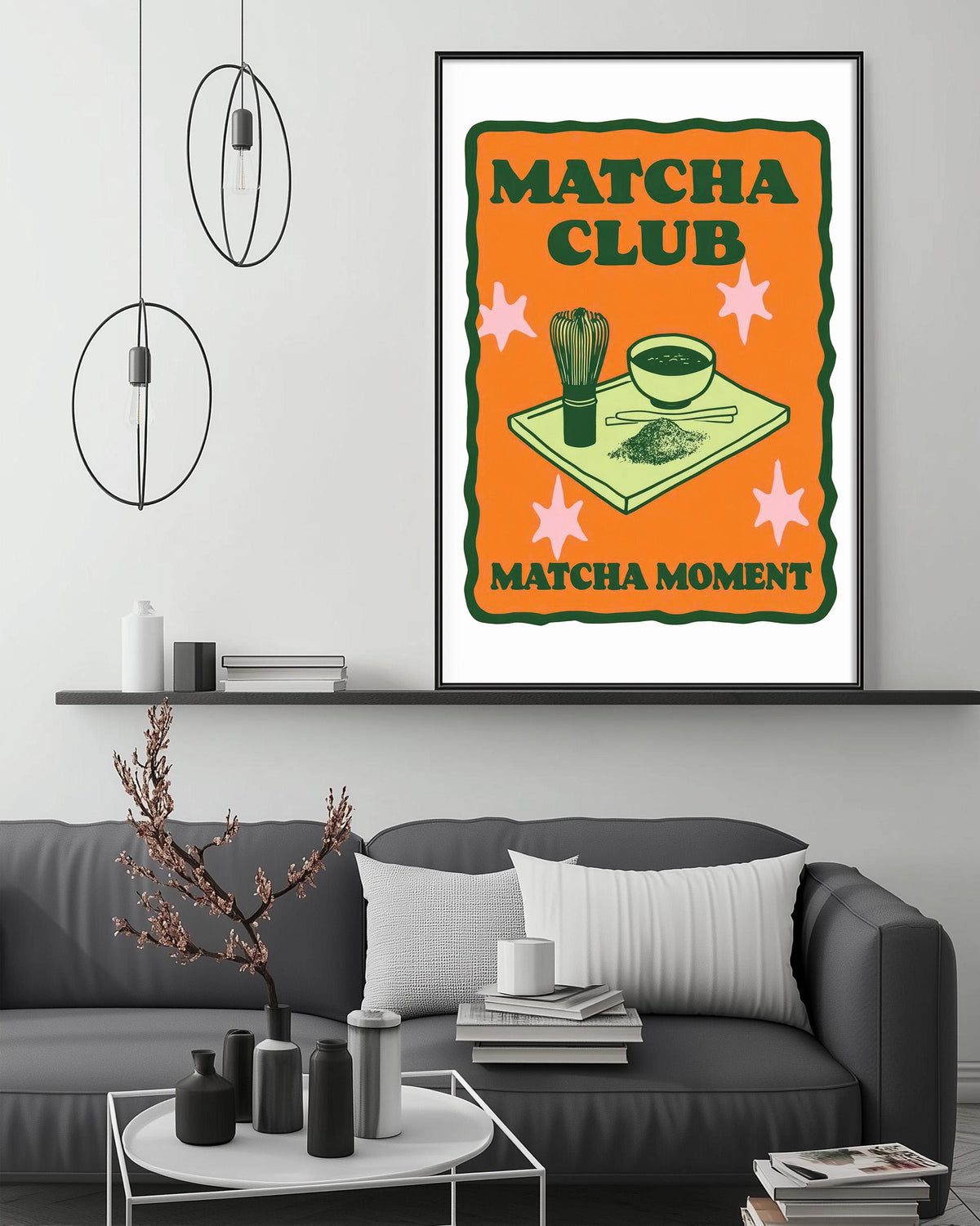 a framed poster with an illustration of a bowl and a cup, along with the text "MATCHA CLUB MATCHA MOMENT" and a star-shaped design. The poster is displayed on a wall above a gray couch, which has a coffee table in front of it. The room has a minimalist and modern design, with a few decorative items and lighting fixtures.