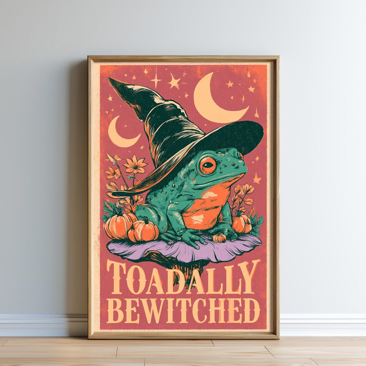 The image is a framed poster featuring a frog wearing a witch's hat, surrounded by autumnal elements such as pumpkins and flowers, with the text "Toadally Bewitched" prominently displayed.