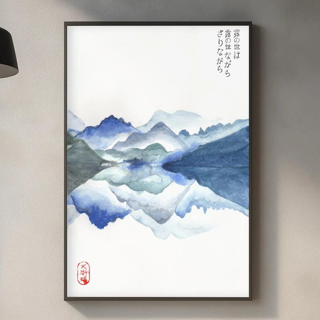 a kitchen counter with a framed watercolor painting of mountains hanging on the wall above it. The painting depicts a serene landscape with blue and white mountains. The kitchen counter also has various items such as a cutting board, a bottle, and a vase.