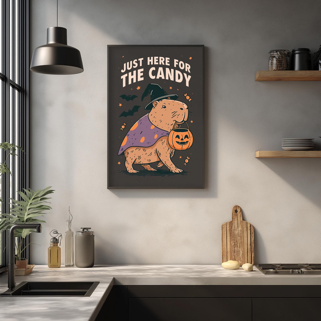 a kitchen counter with various items, including a pumpkin-shaped bowl, a wooden cutting board, and a potted plant. Above the counter, there is a framed poster with a cartoon image of a bear dressed in a witch's hat and holding a pumpkin, along with the text "Just here for the candy".