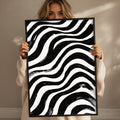 A woman is holding up a large framed print with a striking black and white abstract design featuring wavy lines.