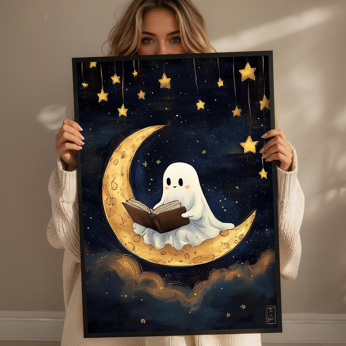 A woman holds up a framed artwork depicting a ghost reading a book while sitting on a crescent moon, surrounded by stars.