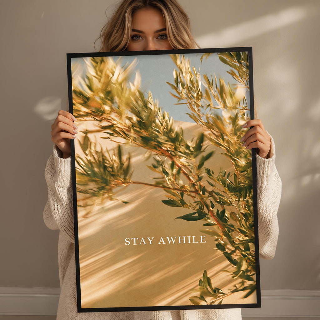 A woman holding a framed print with a blurred image of a tree and the text "STAY AWHILE" on it.
