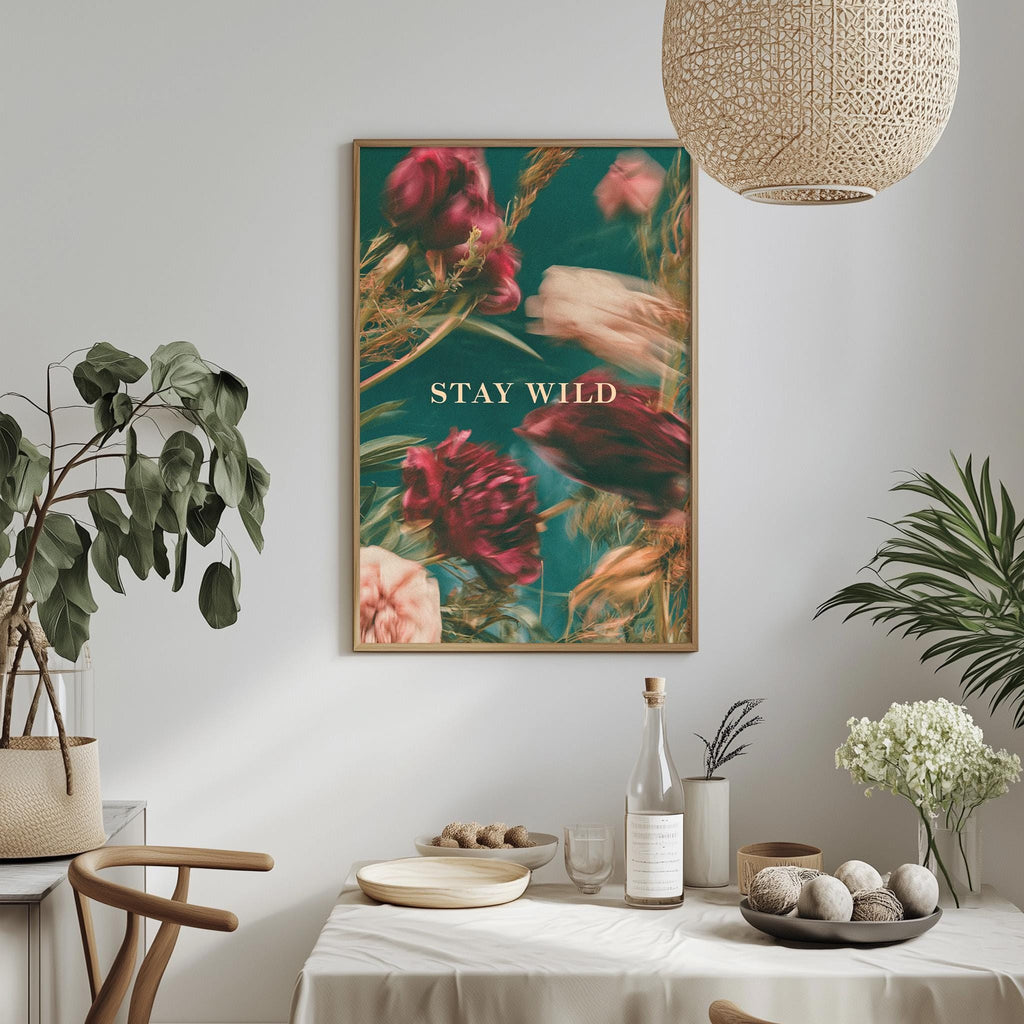a dining table with a white tablecloth, a bowl of fruit, a bottle, and a vase with flowers. Above the table, there is a framed poster with the text "STAY WILD" in the center.