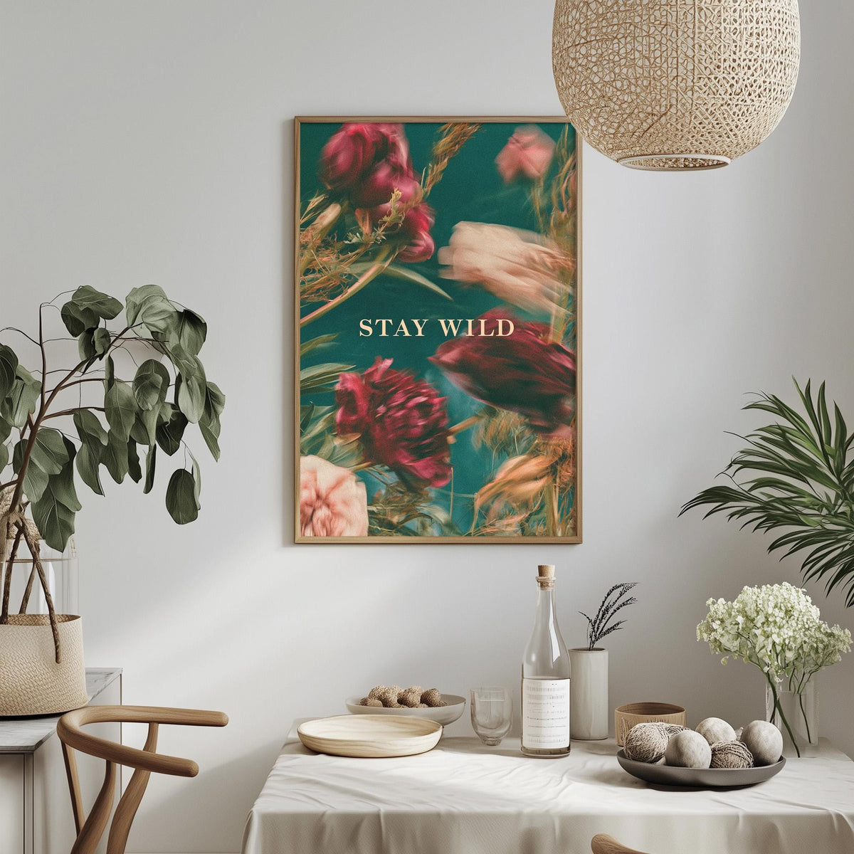 a dining table with a white tablecloth, a bowl of fruit, a bottle, and a vase with flowers. Above the table, there is a framed poster with the text "STAY WILD" in the center.