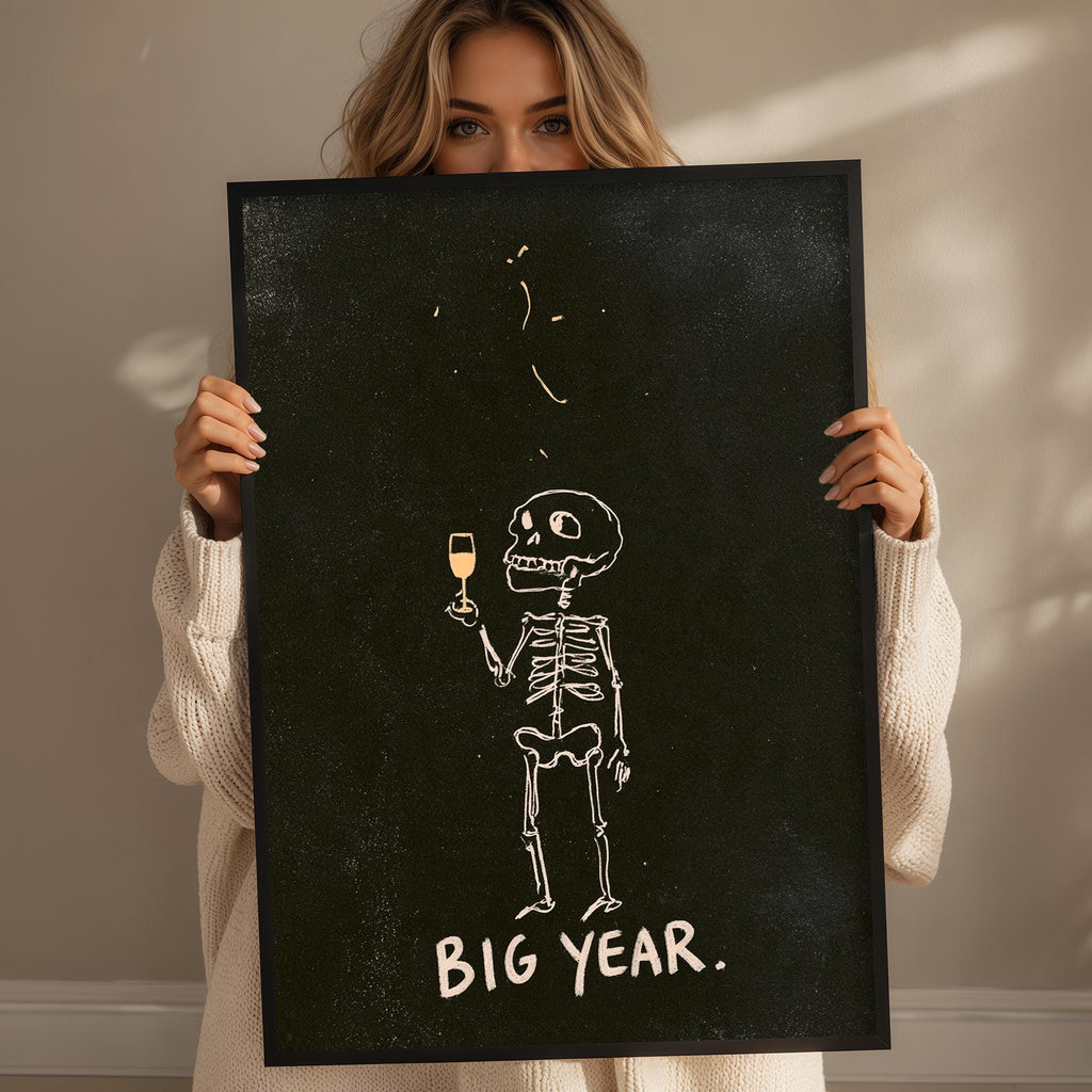 A woman holding a black frame with a skeleton drawing and the text "BIG YEAR."