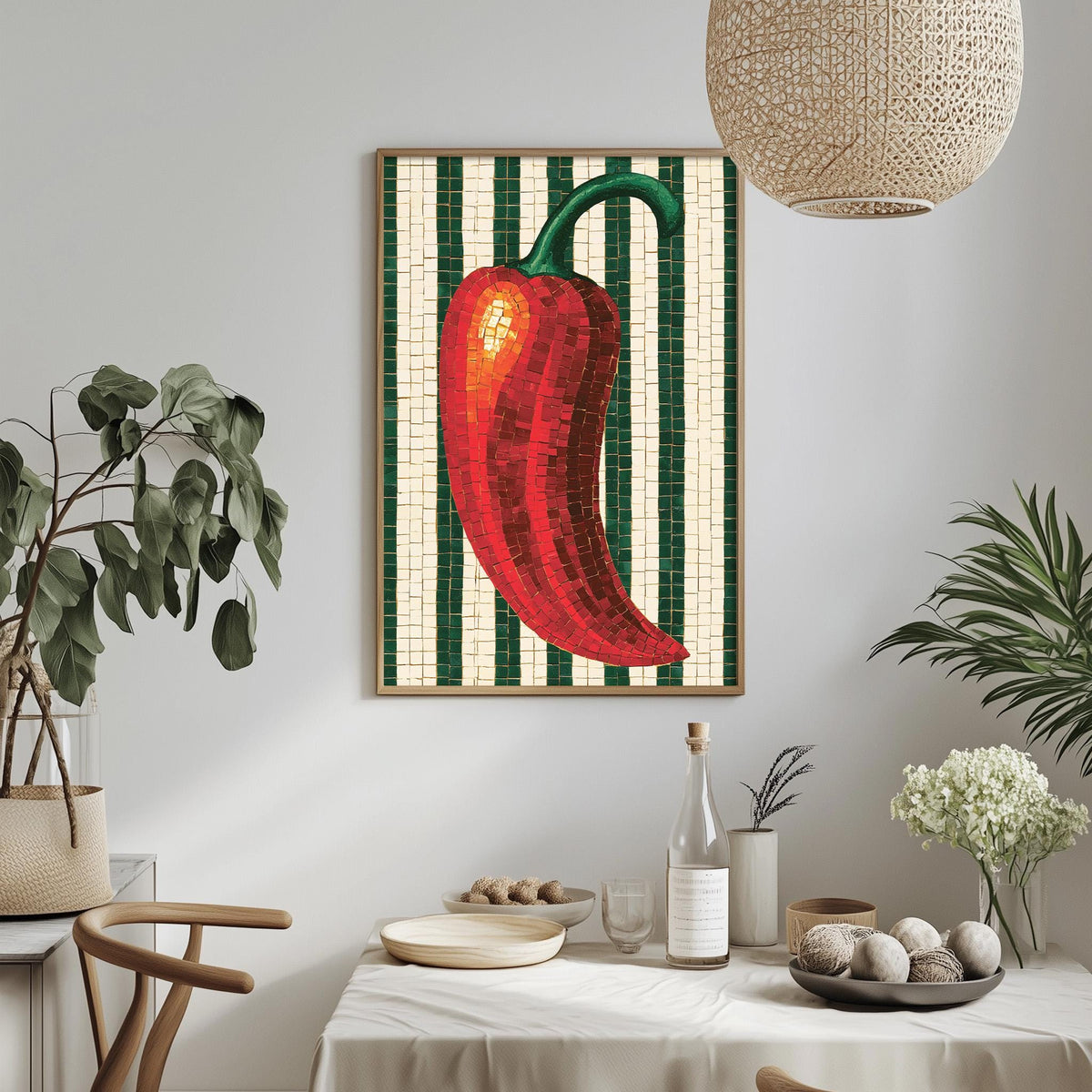a dining table with a white tablecloth, a bowl of fruit, a bottle, and a vase with flowers. Above the table, there is a framed picture of a red pepper on a green and white striped background.