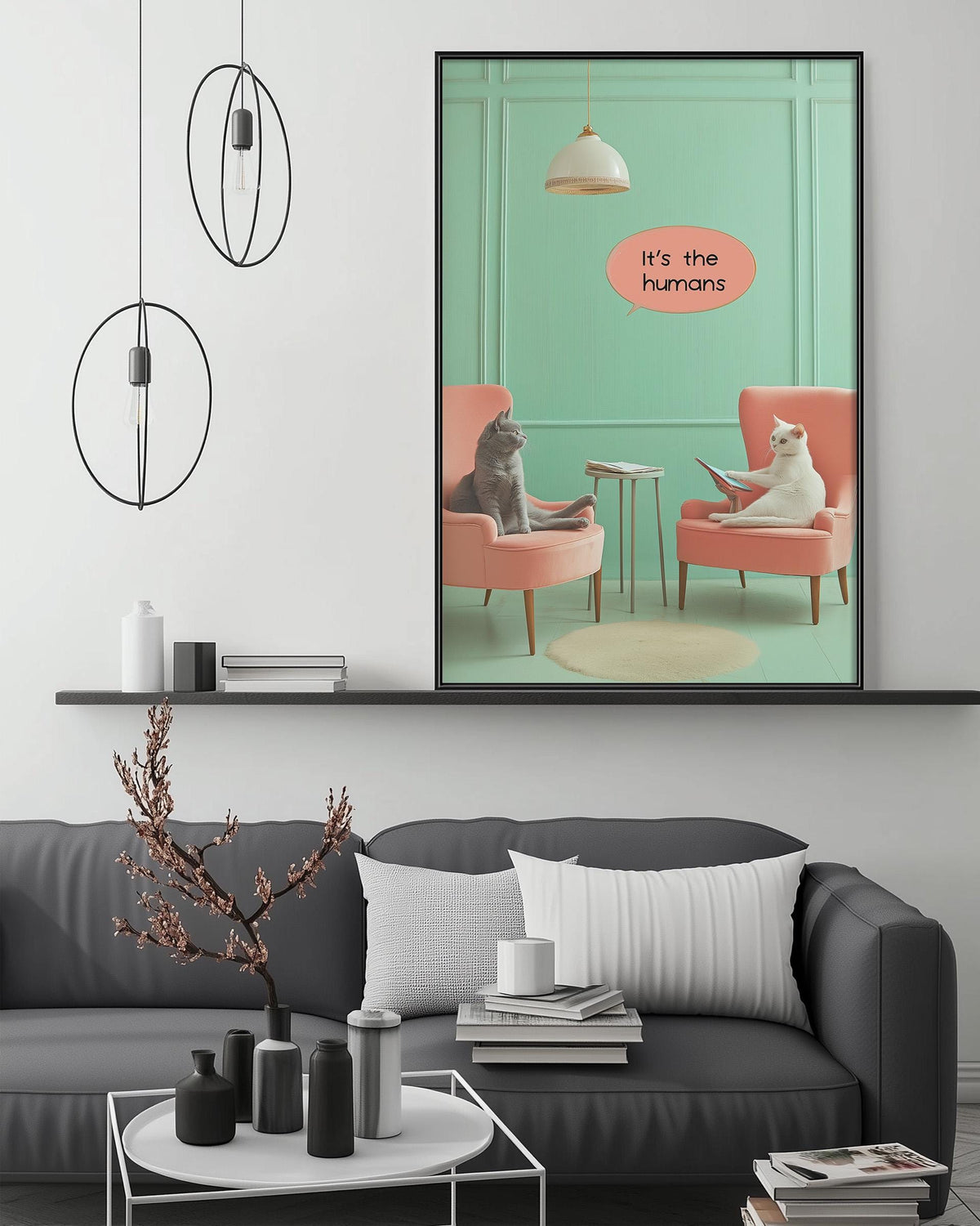 a living room with a gray couch, a coffee table, and a framed poster of two cats sitting in chairs. The poster has a humorous caption that reads "It's the humans".