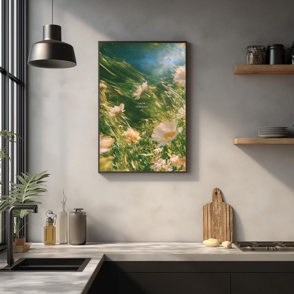 a kitchen counter with a framed picture of flowers hanging above it, a wooden cutting board, and various kitchen items such as a bottle, a cup, and a bowl.