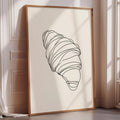 A framed illustration of a croissant is displayed on a wall in a room with a window.