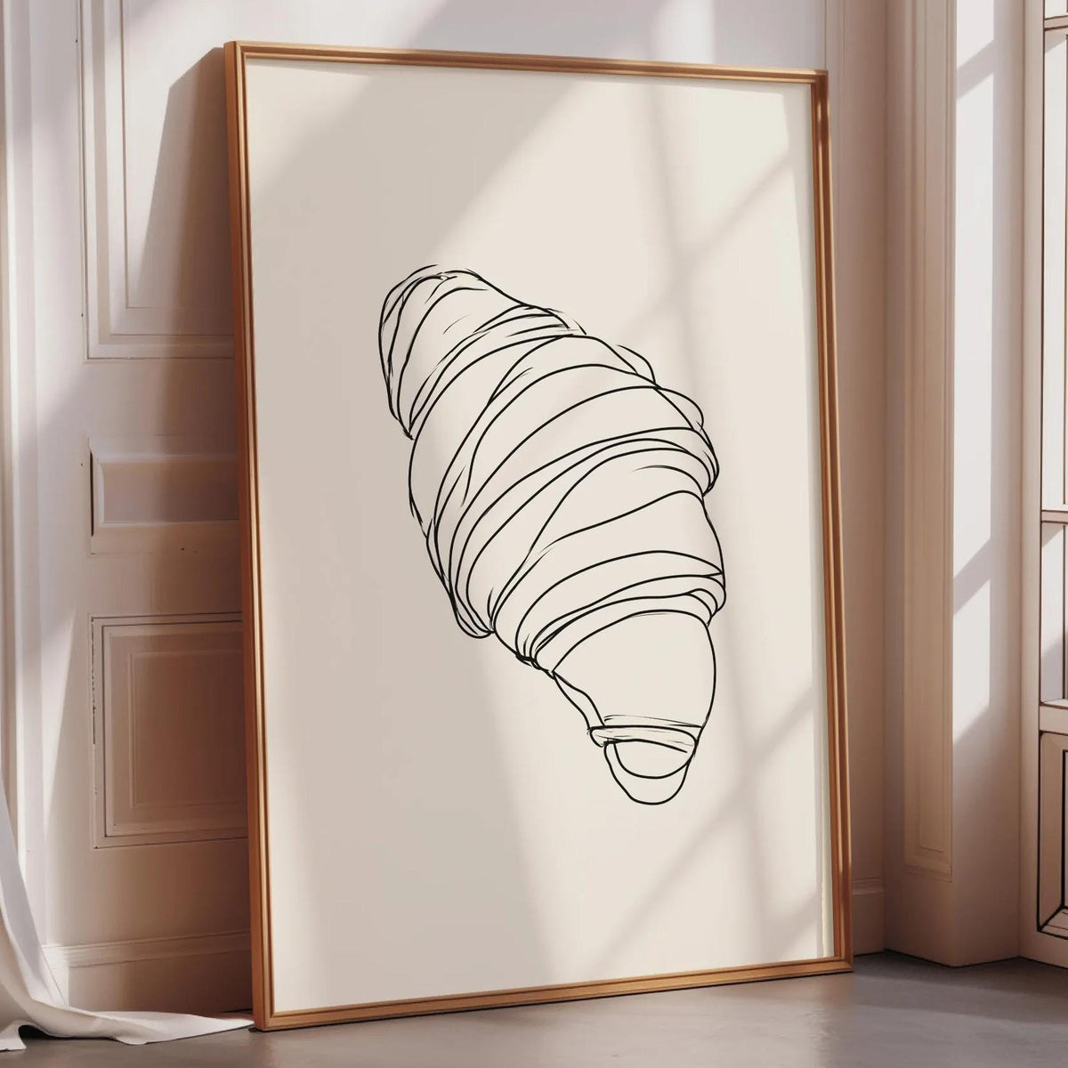 A framed illustration of a croissant is displayed on a wall in a room with a window.