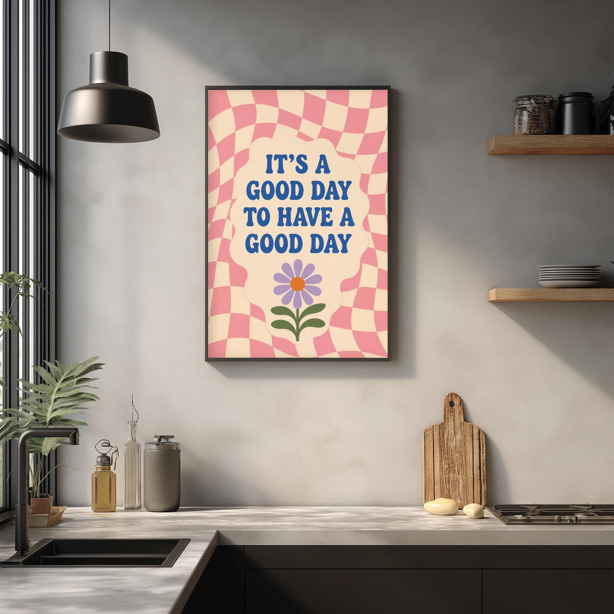 a kitchen counter with a framed poster hanging on the wall above it. The poster has a checkered pattern and a flower design, and it displays the text "It's a good day to have a good day".